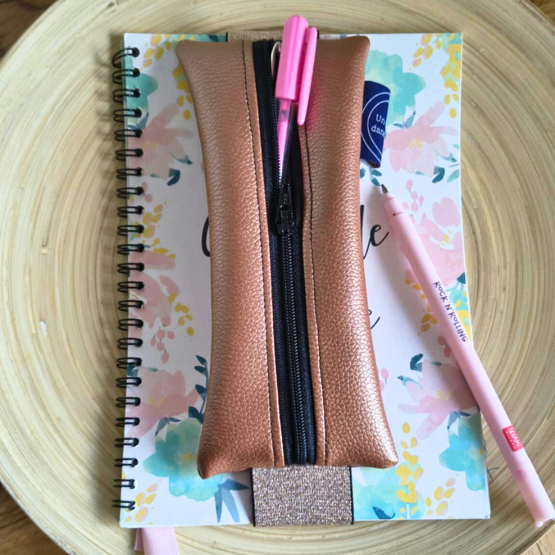 Trousse marque-page bronze en simili cuir, posée sur un carnet fleuri avec un stylo rose, accessoire pratique et élégant pour garder ses stylos à portée de main.