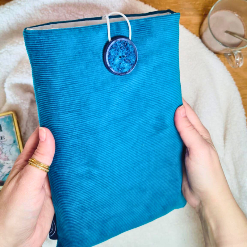 Pochette à livre en tissu crème avec des motifs dorés de citrouilles et feuillages d’automne, fermée par un bouton décoratif, posée sur une table rustique avec des accessoires chaleureux de saison.