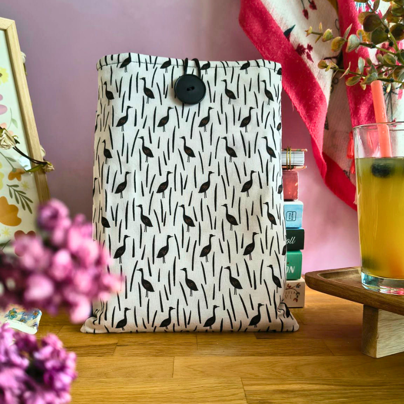 Pochette livre en tissu clair avec motif oiseaux noirs et étui assorti, posée sur une table en bois avec ambiance cosy.