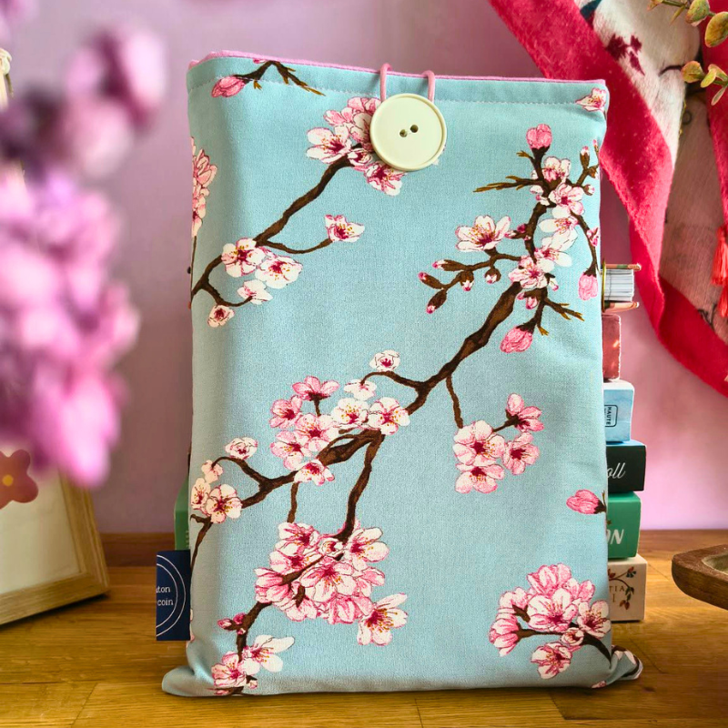 Pochette livre en tissu bleu avec motif cerisier du Japon rose, posée sur une table en bois dans une ambiance déco douce.