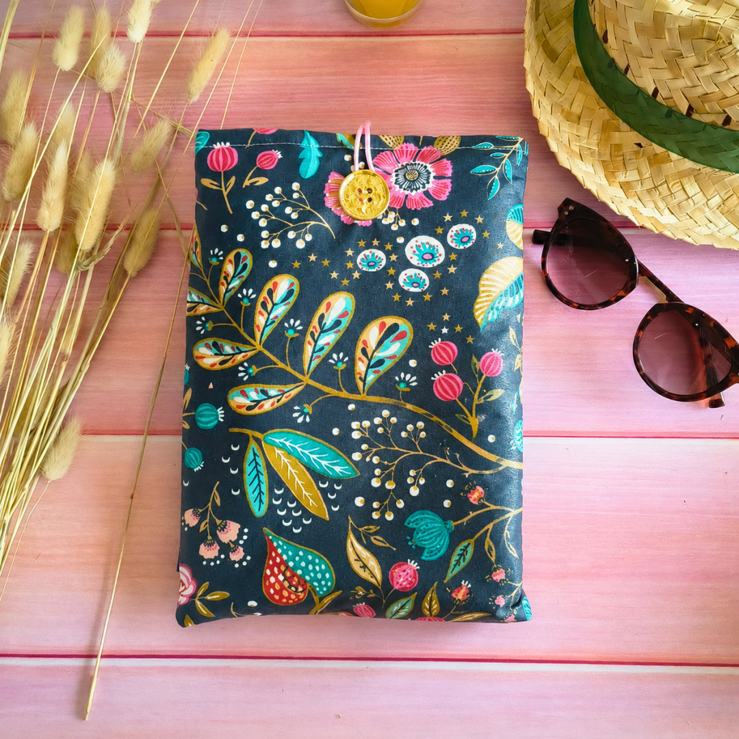 Pochette de livre imperméable à motifs floraux colorés posée sur une table rose avec chapeau de paille, lunettes de soleil et ambiance estivale.