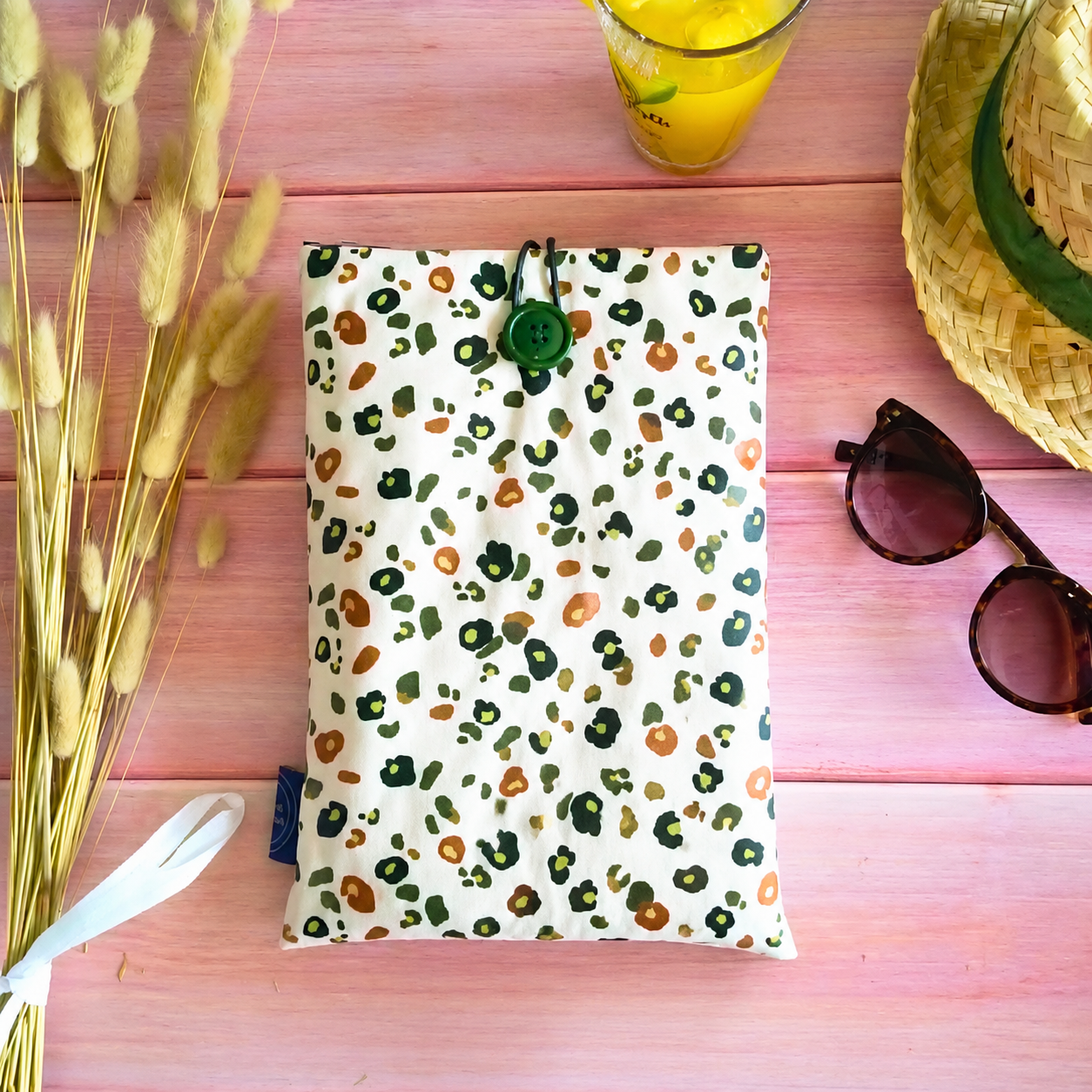 Pochette de livre imperméable à motif léopard vert posée sur une table rose avec lunettes de soleil et chapeau d’été.