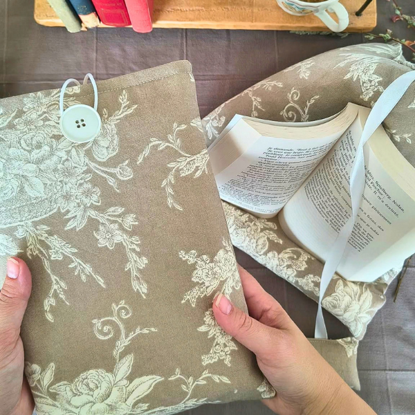 Pochette de livre beige fleurie avec bouton, posée sur une table auprès de livres et d’une tasse